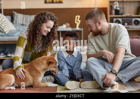 Kleiner schwarzer Junge, der mit lächelnden Adoptiveltern sitzt und mit Shiba Inu Hund auf dem Boden im Kinderzimmer spielt, glückliches Familienkonzept Stockfoto