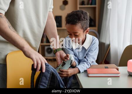 Mittelgroße Aufnahme von Little Black Boy, der Schultasche mit Büchern und Briefpapier packt, während der unerkennbare Adoptivvater dem Sohn hilft, sich auf die Schule vorzubereiten Stockfoto