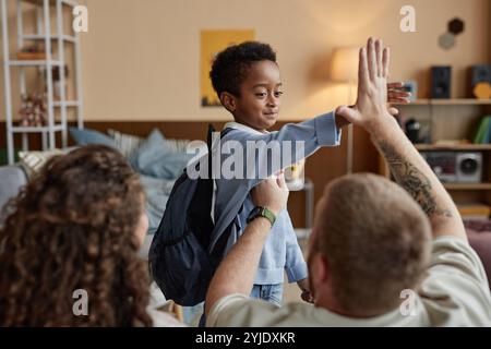 Mittlere Aufnahme des fröhlichen kleinen Schwarzen Jungen, der High Five an unterstützenden Vater gibt, während Eltern das Kind im Kinderzimmer auf die Schule vorbereiten, Happy Adoptive fami Stockfoto