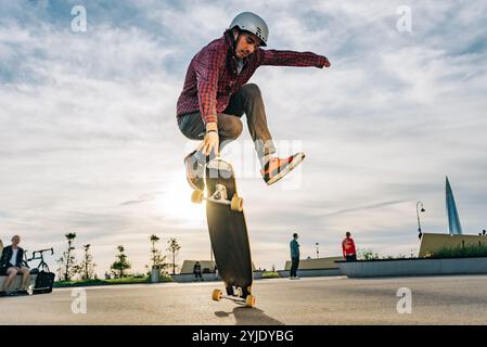 Ein junger Mann in einem Helm springt auf ein Longboard in einem Skatepark, beleuchtet von der untergehenden Sonne, und zeigt seine Fähigkeiten in einer urbanen Stadtlandschaft Stockfoto