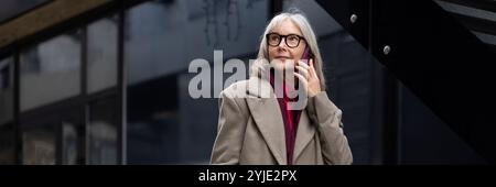 Eine ältere Frau mit Brille spricht auf ihrem Smartphone, während sie vor einem modernen Gebäude in einem Stadtgebiet steht Stockfoto