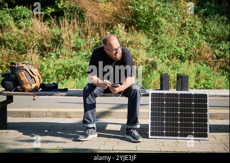 Bärtiger Mann mit Smartphone, das mit Photovoltaik-Sonnenkollektor verbunden ist. Integration nachhaltiger erneuerbarer Energien in den Alltag, demonstrieren die praktische Nutzung von Solarstrom für Ladegeräte. Stockfoto