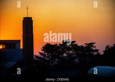 Die Silhouette eines Kirchturms und Bäume vor einem lebendigen Sonnenuntergangshimmel unterstreicht die friedliche und ruhige Abendatmosphäre in einer urbanen Umgebung. Stockfoto