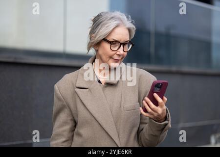 Eine ältere Geschäftsfrau überprüft ihr Smartphone, während sie an einem sonnigen Nachmittag vor einem modernen Bürogebäude steht Stockfoto