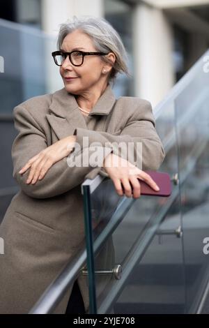 Eine ältere Frau in stilvoller Brille lehnt sich an das Glasgeländer und genießt einen sonnigen Tag in einer städtischen Umgebung Stockfoto