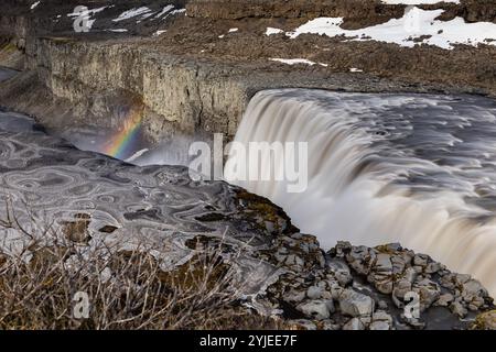 Regenbogen über Dettifoss Wasserfall im Vatnajokull Nationalpark im Nordosten Islands, Blick auf Wasserfall und Canyon mit Basaltsäulen, Eis und Schnee. Stockfoto