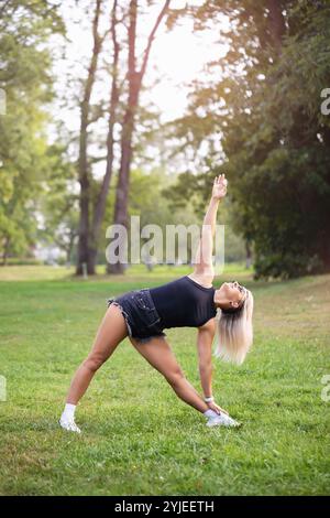 Sportliche Frau mit europäischem Aussehen, Trikonasana-Übungen, Dreiecksposen, in kurzen Shorts und Tank-Top im Park an sonnigen warmen Abend Stockfoto