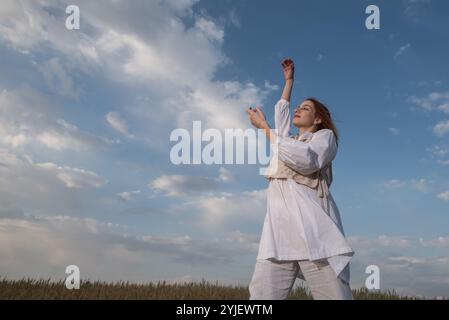 Eine junge Frau in weißem Kleid, die draußen vor blauem, bewölktem Himmel tanzt. Stockfoto