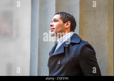 Ein Mann in einem dunklen Mantel blickt nach oben vor einem Betonhintergrund. Sanfte Beleuchtung unterstreicht seinen kontemplativen Ausdruck und die Textur seines Mantels. Stockfoto
