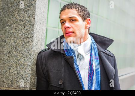Eine Person mit kurzen lockigen Haaren lehnt sich an eine strukturierte Wand, trägt einen dunklen Mantel und einen blauen karierten Schal. Der Hintergrund ist grün, aus Glas... Stockfoto