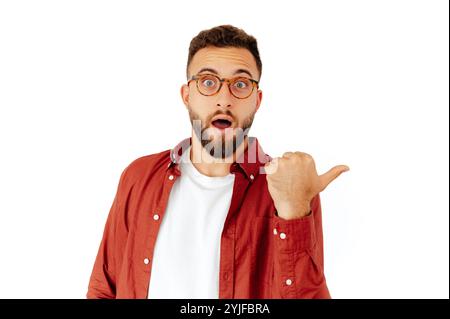 Attraktiver aufgeregter kaukasischer oder arabischer junger Mann in einem roten Hemd mit Brille, sieht inspiriert und überrascht aus, während er auf isoliertem weißem Hintergrund steht und den Finger seitlich zu einem Kopierraum zeigt Stockfoto