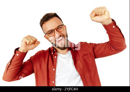 Glücklicher junger arabischer oder kaukasischer Mann in Brille, der mit erhobenen Fäusten feiert, Aufregung und Begeisterung zeigt, in einem roten Hemd gekleidet, auf isoliertem weißem Hintergrund stehend Stockfoto