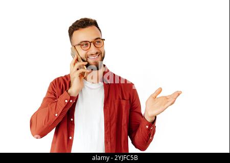 Ein fröhlicher bärtiger Mann in Brille, der ein rotes Hemd und ein weißes T-Shirt trägt, mit einem freundlichen Lächeln am Telefon spricht und mit der Hand gestickt, isoliert auf weißem Hintergrund Stockfoto