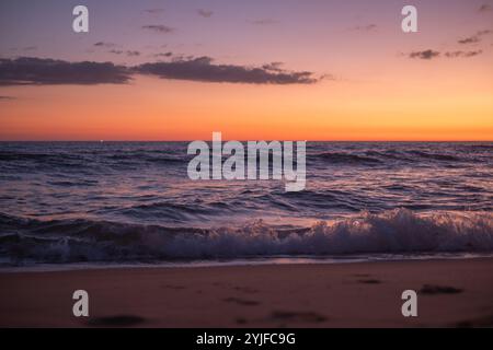 Die Sonne untergeht über dem Meer und färbt den Himmel mit leuchtenden Orange- und Violetttönen, während sanfte Wellen gegen die Sandküste schlängeln und eine Ruhe schaffen Stockfoto