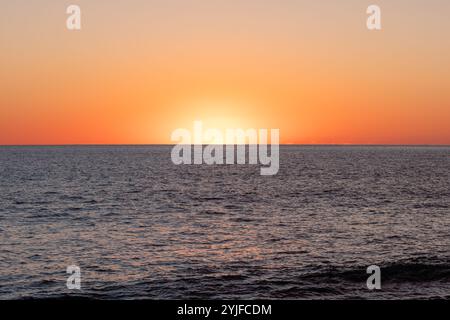 Goldene und orangene Farben verschmelzen im Himmel, während die Sonne über dem ruhigen Meer untergeht, und bieten einen ruhigen und malerischen Blick auf die Küste am Abend. Stockfoto