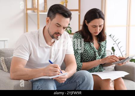 Eine junge Frau, die Berechnungen macht, während ihr Mann zu Hause etwas aufschreibt. Insolvenzkonzept Stockfoto