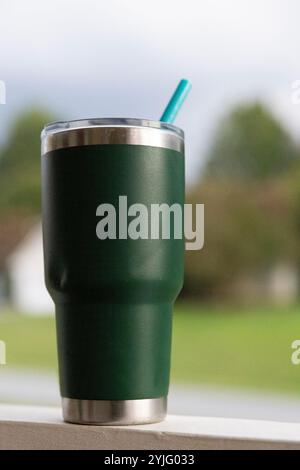 Nahaufnahme eines grünen Reisebechers aus Edelstahl mit blauem Silikonstroh auf dem Geländer der Veranda, perfekt für den morgendlichen Eiskaffee oder zum Trinken Ihres W Stockfoto