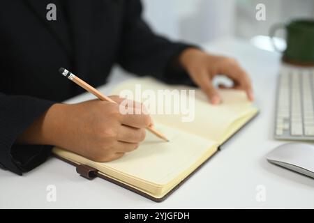 Nicht erkennbare Geschäftsfrau, die in ein Tagebuch schreibt, um sich an eine Aufgabe am Schreibtisch zu erinnern Stockfoto