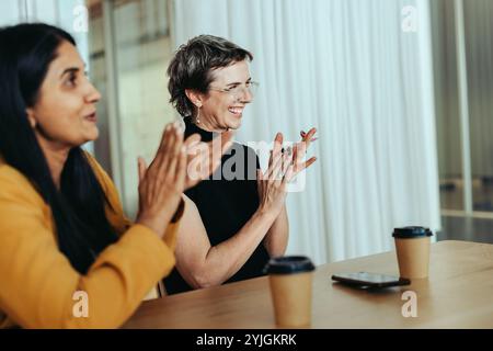 Ein fröhliches Büromeeting mit zwei klatschenden und lächelnden Personen, das eine einnehmende und positive Atmosphäre schafft. Kaffeetassen sind auf dem Tisch sichtbar, en Stockfoto