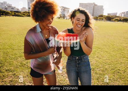 Zwei College-Freunde teilen sich Lachen und ein Stück Wassermelone in einem urbanen Park, um die Freude und Spontaneität der Freundschaft an einem sonnigen Tag festzuhalten. Stockfoto