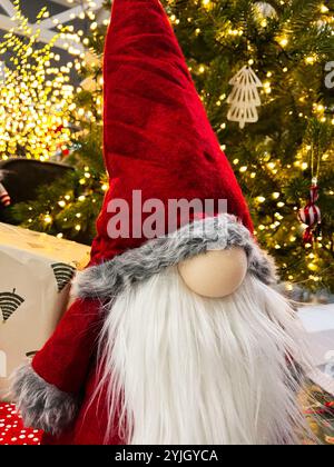 Ein bezaubernder Weihnachtszwerg mit rotem Hut und langem Bart steht neben einem geschmückten Weihnachtsbaum, umgeben von funkelnden Lichtern und gewickelten Geschenken. Stockfoto
