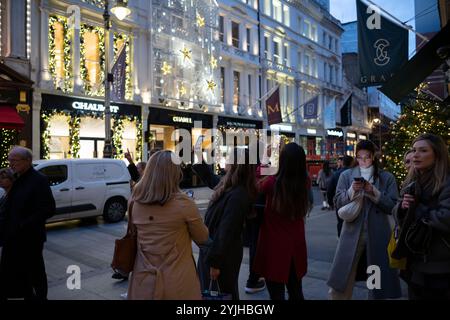 Touristen halten an und machen Fotos von den Weihnachtsfenstern und Weihnachtslichtern entlang der Old Bond Street, Mayfair, im Zentrum von London, England, Großbritannien Stockfoto