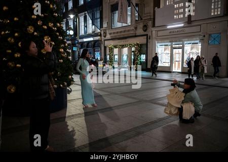 Touristen halten an und machen Fotos von den Weihnachtsfenstern und Weihnachtslichtern entlang der Old Bond Street, Mayfair, im Zentrum von London, England, Großbritannien Stockfoto