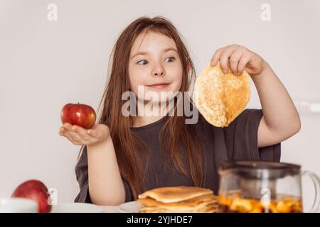 Ein kleines Teenager-Mädchen, das einen Apfel in der einen Hand in der anderen Pfannkuchen hält. Das Mädchen wählt zwischen einem Apfel und einem Pfannkuchen zum Frühstück, sieht aus, als ob es ihn leckt Stockfoto