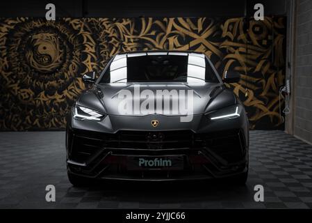 Ein Lamborghini Urus Performante mit mattschwarzer Oberfläche wird in Innenräumen ausgestellt. LED-Scheinwerfer und -Embleme sind sichtbar und stehen auf einer goldenen und schwarzen Wandmalerei Stockfoto