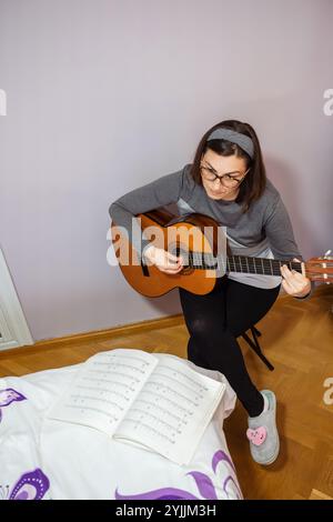 Die Musikerin spielt zu Hause klassische Gitarre, umgeben von Musiknoten, voll in das Lernen und die Perfektion ihres Handwerks eingetaucht und kreiert wunderschöne Melodien, die den Raum mit gefühlvollen Klängen füllen Stockfoto