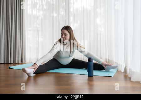 Gen Z schwangere Frau praktiziert Yoga zu Hause mit einer Wasserflasche in der Nähe. Werdende Mutter im pränatalen Trainingskurs. Stockfoto