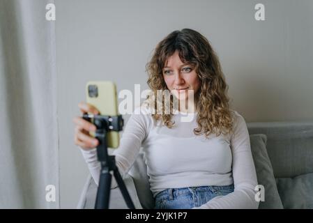 Die junge Influencerin erstellt Inhalte von zu Hause aus, sitzt auf einem Sofa mit einem Smartphone auf einem Stativ und interagiert mit ihrem Publikum über soziale Medien Stockfoto