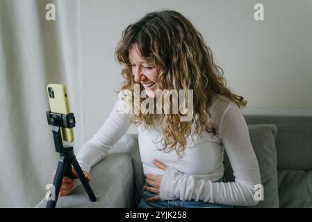 Die junge, lockhaarige Inhaltserstellerin lacht, während sie zu Hause mit einem Smartphone auf einem Stativ einen lustigen Vlog aufnimmt und so ansprechende Inhalte für ihr Online-Publikum erstellt Stockfoto