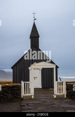 Schwarze Holzkirche mit einem Kreuz auf dem Turm. Weiße Details. Auf einer Weide, in der Nähe des Ozeans, auf einer Halbinsel gelegen. Budakirkja, Island. Stockfoto