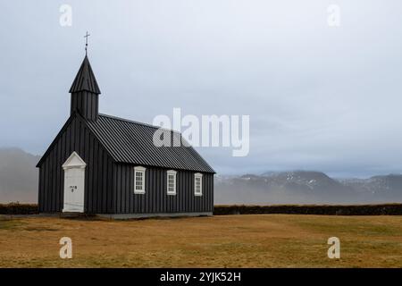 Schwarze Holzkirche mit einem Kreuz auf dem Turm. Weiße Details. Auf einer Weide, in der Nähe des Ozeans, auf einer Halbinsel gelegen. Budakirkja, Island. Stockfoto