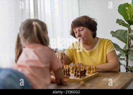 Ein fröhliches Mädchen spielt Schach mit ihrer Großmutter am Tisch und schafft wertvolle Familienerinnerungen. Hochwertige Fotos Stockfoto