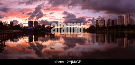 Der dramatische orangene Himmel spiegelt sich im ruhigen Wasser des Sees wider, während die Silhouetten von Gebäuden und Bäumen in der Abenddämmerung eine urbane Umgebung schaffen. Stockfoto