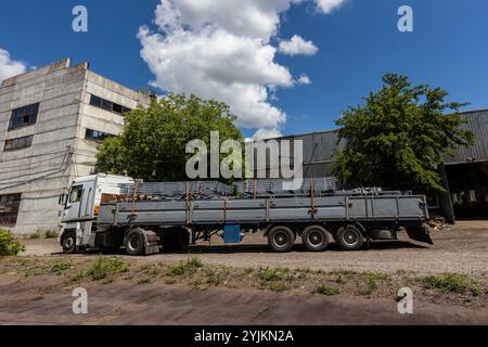 An einem sonnigen Tag steht ein großer Tieflader mit Metallträgern beladen in der Nähe von Industriegebäuden. Bäume und blauer Himmel mit Wolken sind im Ba sichtbar Stockfoto