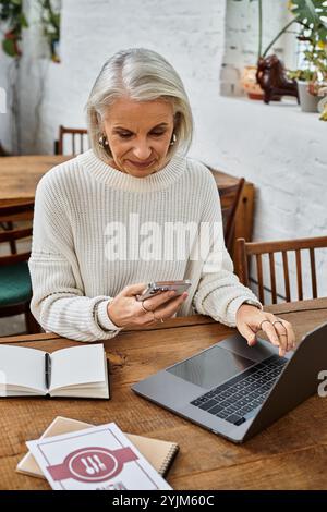 Eine schöne grauhaarige Frau multitasst in einem rustikalen Café auf ihrem Smartphone und Laptop. Stockfoto