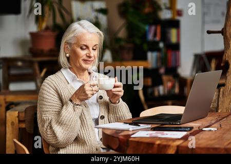 Eine schöne grauhaarige Frau sitzt an einem Holztisch und lächelt beim Kaffeetrinken. Stockfoto