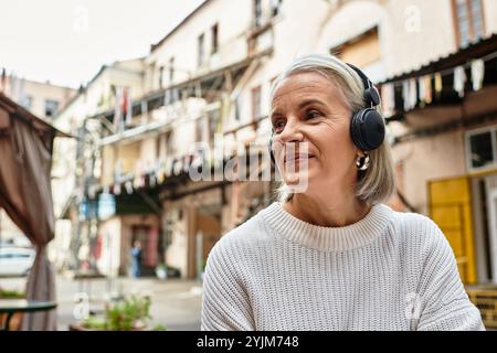 Eine schicke ältere Frau mit grauen Haaren genießt Musik, während sie sich im Freien in einer gemütlichen Umgebung entspannt. Stockfoto