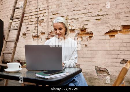 Eine schöne Frau mit grauem Haar sitzt an einem Tisch, in ihren Laptop getaucht und schlürft Kaffee. Stockfoto