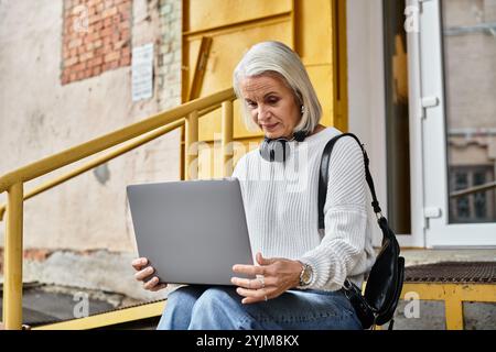 Eine reife Frau mit grauen Haaren arbeitet mit ihrem Laptop, während sie auf einer hellen Treppe ruht. Stockfoto
