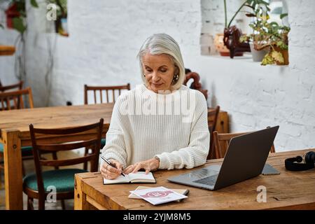 Eine reife Frau mit grauen Haaren sitzt an einem Holztisch und konzentriert sich auf das Schreiben in ihrem Notizbuch. Stockfoto