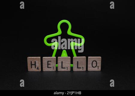 Fluoreszierender grüner Plastikmann, der hinter den Scrabble Holzbuchstaben-Fliesen mit HALLO-Wort auf schwarzem Hintergrund steht, Welt-Hallo-Tag Stockfoto