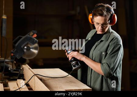 Eine gelernte Schreinerin konzentriert sich intensiv, während sie Elektrowerkzeuge für ein Holzprojekt in ihrem Werkstattbereich verwendet. Stockfoto