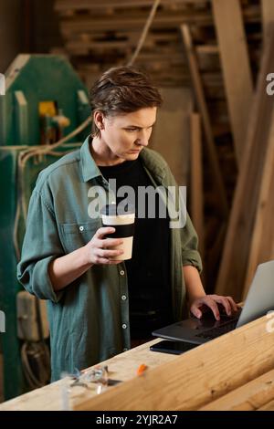 Eine gelernte Schreinerin konzentriert sich auf ihren Laptop, Kaffee in der Hand, in einer geschäftigen Werkstatt voller Holzprojekte. Stockfoto