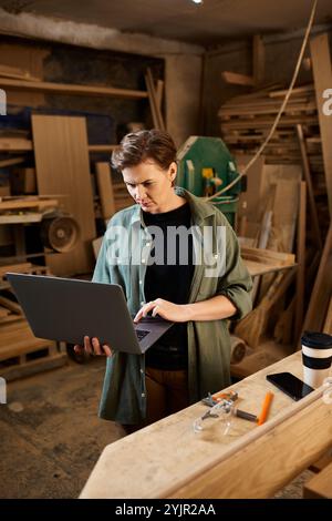 Ein erfahrener Zimmermann bewertet Pläne auf einem Laptop in einer lebendigen Werkstatt voller Holzkreationen und Werkzeuge. Stockfoto