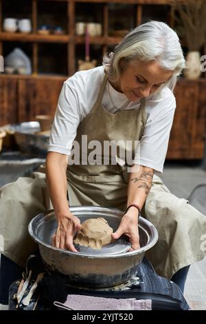 Eine reife Frau mit silbernem Haar konzentriert sich intensiv, während sie Ton auf einem Keramikrad formt. Stockfoto