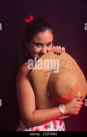 Schöne Frau, die seitlich steht und in die Kamera blickt, in einem bunten Kleid, das einen Hut mit der Hand hält. Isoliert auf farbenfrohen rauchigen Hintergrund. Stockfoto